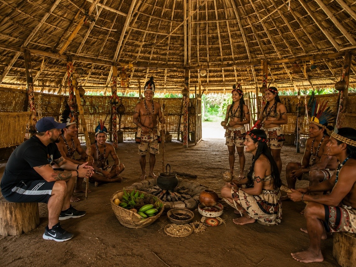 Visitante y miembros de una comunidad indígena reunidos dentro de una maloca tradicional de techo de paja